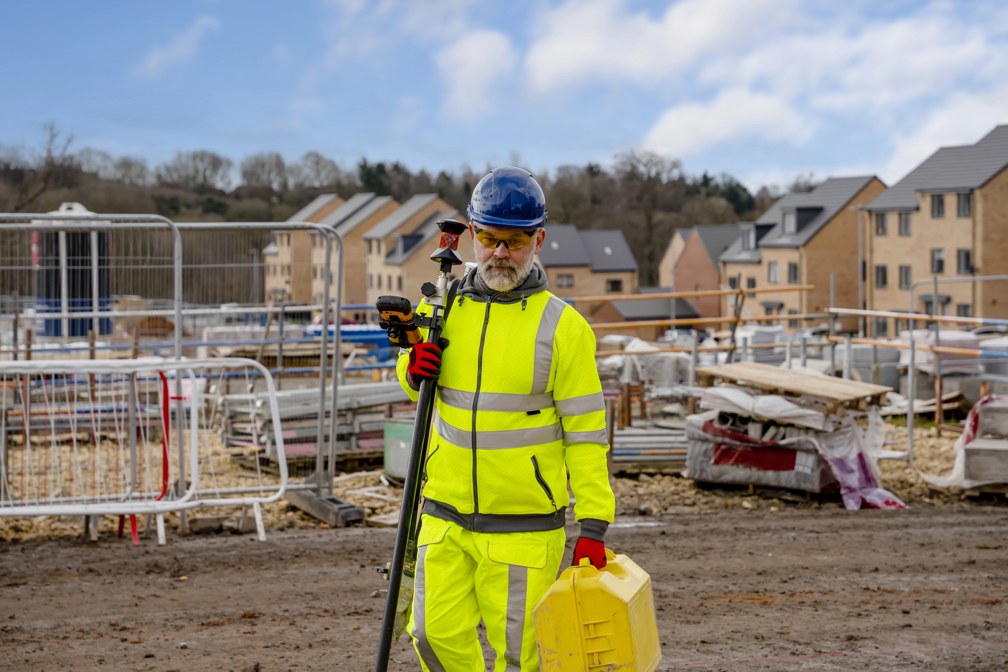 Older bearded construction worker in bright high visibility jacket and pants, carrying equipment in a yellow case at an apartment building construction site